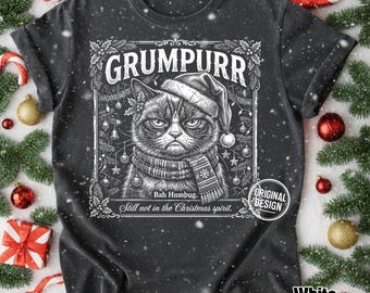 Może przedstawiać: Ciemnoszary t-shirt z białą grafiką przedstawiającą kota w czapce Mikołaja i szaliku. Napis głosi GRUMPURR, Bah Humbug oraz Still not in the Christmas spirit. Wzór jest otoczony gałązkami sosny i świątecznymi ozdobami.