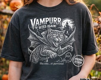 Może przedstawiać: Ciemnoszary t-shirt z białą grafiką przedstawiającą kota ze skrzydłami nietoperza, kłami i peleryną. Napis głosi VAMPURR BITES AGAIN! oraz This time... it's purrrsonal. Widoczny jest zamek, błyskawice oraz nagrobek z napisem R.I.P.