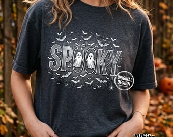 Może przedstawiać: Ciemnoszary melanżowy t-shirt z białym nadrukiem graficznym przedstawiającym słowo SPOOKY dużymi literami. Wzór zawiera dwa duszki, kokardę, pajęczynę i liczne małe nietoperze. Okrągła odznaka zawiera napis ORIGINAL DESIGN.