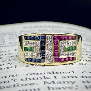 Puede incluir: Un anillo de oro con un diseño geométrico. El anillo presenta piedras preciosas de corte cuadrado en azul, rojo, verde y blanco, dispuestas en un patrón rectangular. El anillo está sobre un libro.