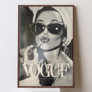 May include: Black and white framed print of a woman applying lipstick, wearing sunglasses and a towel on her head. The word "VOGUE" is displayed in large letters at the bottom of the image. The artwork has a vintage, glamorous aesthetic.