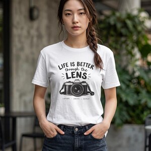 May include: A white short-sleeved t-shirt featuring a black graphic of a camera and the text "LIFE IS BETTER through the LENS" along with the words "CAPTURE CREATE REPEAT" printed in black ink on the front center of the garment.