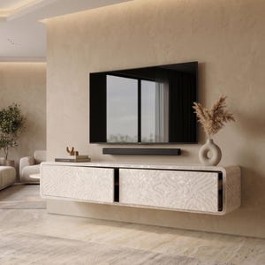May include: A modern living room setup featuring a large flat-screen TV mounted on a beige wall. Below the TV is a white floating cabinet with two drawers, adorned with a patterned design. A soundbar is positioned below the TV. A vase with dried plants sits on the cabinet.