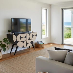 May include: A modern black and beige cabinet with a geometric design, holding a flat-screen TV. The cabinet has angled legs and is set against a white wall. A woven basket and a monstera plant are nearby.