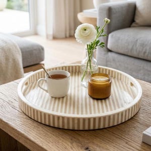 Könnte beinhalten: Ein runder Dekoteller mit Streifenmuster trägt eine Tasse Tee mit einem Löffel, eine kleine Glasvase mit einer weißen Blume und eine Kerze in einem braunen Glas. Der Teller steht auf einem Holztisch.