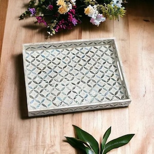 May include: Rectangular decorative tray with a white frame and a geometric pattern of overlapping circles. The tray's surface has a mother-of-pearl effect, and the design is in shades of gray and blue. The tray is on a wooden surface.