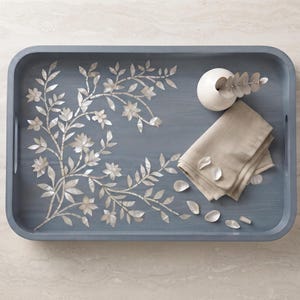 Puede incluir: Una bandeja de servicio rectangular de color azul pizarra con asas laterales integradas, que presenta un patrón floral y de enredaderas con incrustaciones de nácar. Un pequeño jarrón de cerámica blanca con una rama de eucalipto y una servilleta de lino beige.