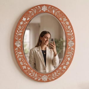 May include: An oval wall mirror featuring a terracotta-colored frame decorated with intricate white floral and vine inlays made of mother-of-pearl. The reflective glass surface is smooth and clear, framed by the ornate, symmetrical botanical pattern.