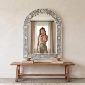 May include: An arched mirror with a floral design border in shades of blue, white, and gray. The mirror is above a wooden bench. The reflection shows a person taking a photo.
