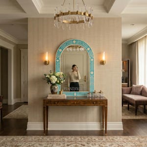 May include: An arched wall mirror with a bright turquoise frame featuring white floral and leaf inlays. The mirror is positioned above a rectangular wooden console table with three drawers, holding a glass vase of flowers and small glass jars.