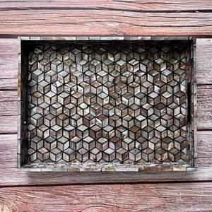 May include: Rectangular decorative tray with a geometric pattern. The tray's surface is covered in small, iridescent, cube-shaped tiles in shades of brown, beige, and gray. The tray has a dark border and integrated handles.