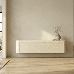 Könnte beinhalten: Ein cremefarbener, an der Wand befestigter Schrank mit einem geschnitzten Blumendesign. Obenauf steht eine kleine, beige Vase mit getrockneten Zweigen sowie ein kleines, rundes, beiges Dekorationsobjekt. Der Schrank befindet sich in einem Raum mit hellen Wänden und Holzboden.