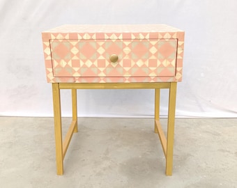 Mesita de noche rosa palo con incrustaciones geométricas de hueso, patas de metal dorado, mesa auxiliar de lujo hecha a mano.