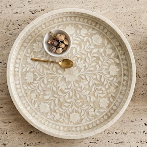 May include: A round cream-colored tray featuring an intricate inlaid floral pattern with white petals and leaves. A small white bowl containing dried pinecones and a gold-toned metal spoon are placed on the surface of the decorative tray.