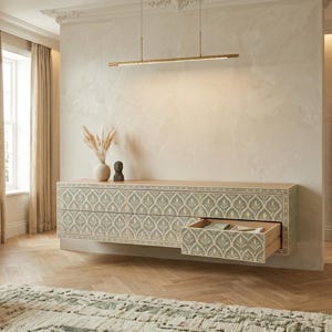 May include: A light wood floating cabinet with a decorative leaf pattern in a neutral color palette. One drawer is open, revealing the interior. The room features a neutral color scheme, a rug, and a gold-toned light fixture.
