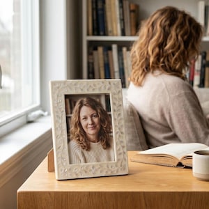 May include: A cream-colored rectangular picture frame with a floral design. The frame holds a portrait of a woman with curly hair. The frame is on a wooden table, with a window and a bookshelf in the background.