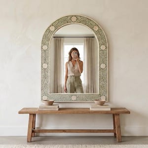 Può includere: Uno specchio da parete ad arco con una cornice verde salvia e un intricato motivo a intarsio con fiori e viti bianche. Lo specchio presenta una parte superiore arrotondata e una base rettangolare, riflettendo una stanza con tende e una panca.