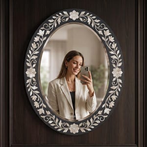 May include: An oval wall mirror featuring a dark frame with an inlaid mother-of-pearl floral vine pattern. The mirror has a reflective glass surface and is mounted on a dark wood-toned wall. The word Handique is printed in small white text.