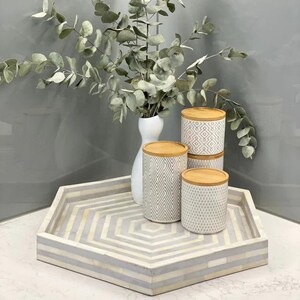 May include: A decorative tray with a geometric pattern holds three patterned canisters with wooden lids. A white vase with eucalyptus branches is behind the canisters. The tray is a light gray and white color.