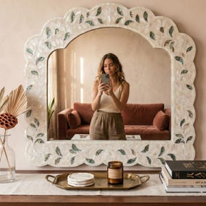 May include: A large, ornate white mirror with a scalloped edge and inlaid leaf designs. The mirror reflects a room with a brown sofa and a person taking a photo. A tray with a candle and coasters sits on a wooden surface.