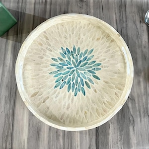May include: Round decorative tray with a cream-colored mosaic pattern and a central blue floral design. The tray is made of a mosaic material and is set on a wooden surface. The tray is a decorative item.