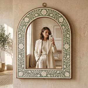 May include: An arched wall mirror featuring a sage green frame with intricate white floral and vine carvings. The mirror has a flat base and a rounded top with a small metal hanging loop. The reflective surface shows a neutral interior space.