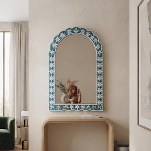 Op de afbeelding: Een boogvormige wandspiegel met een geschulpte lijst in een diepblauwe afwerking en een herhalend wit bloemenpatroon. De spiegel heeft een reflecterend glazen oppervlak en een opvallende gebogen bovenrand voor een helder zicht.