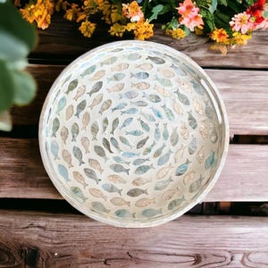 May include: A round decorative tray with a mosaic fish design. The tray features a white background with numerous fish in shades of blue, gray, and beige. The tray is made of glass and is suitable for serving or display.