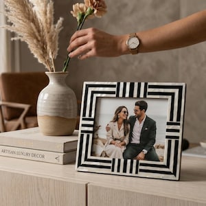 Puede incluir: Un marco de fotos decorativo con un diseño geométrico en blanco y negro. El marco contiene una fotografía de una pareja. También se ve un jarrón con flores secas y una pila de libros.