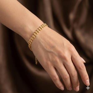 Könnte beinhalten: Ein goldfarbenes Armband mit einem blattartigen Design. Das Armband besteht aus kleinen, verbundenen Segmenten, die ein strukturiertes Aussehen erzeugen. Das Armband wird an einem Handgelenk vor braunem Hintergrund gezeigt.