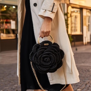 May include: A black, rose-shaped handbag with a gold chain strap and gold handle rings. The bag is held by a person wearing a cream-colored trench coat and a black dress. The background shows a street scene.
