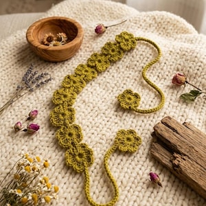 Puede incluir: Una diadema de ganchillo verde oliva hecha a mano y dos accesorios para el cabello a juego con forma de flor. Los accesorios florales están dispuestos sobre una manta de punto color crema, con flores secas y un cuenco de madera en el fondo.