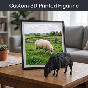 Puede incluir: Una figura negra impresa en 3D de una oveja está sobre una mesa de madera, junto a una foto enmarcada de una oveja pastando en un campo. La foto está en un marco negro. El texto "Custom 3D Printed Figurine" está en la parte superior.