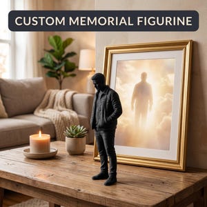 Könnte beinhalten: Schwarze, individuelle Gedenkfigur einer Person, die auf einem Holztisch steht. Die Figur steht neben einem gerahmten Bild mit goldenem Rahmen. Der Text "CUSTOM MEMORIAL FIGURINE" befindet sich oben im Bild.