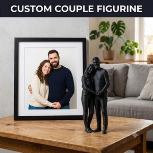 Peut inclure: Une figurine de couple personnalisée en noir mat, représentant deux personnes debout côte à côte enlacées, placée à côté d'une photographie encadrée du même couple avec un passe-partout blanc et un cadre noir, marquée CUSTOM COUPLE FIGURINE.