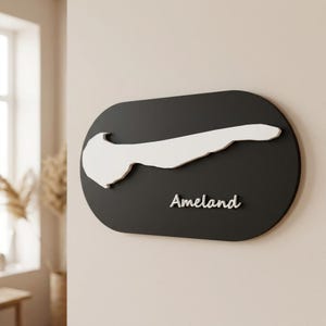 Op de afbeelding: Een zwart ovaal wandbord met een verhoogd wit silhouet van het eiland Ameland. Het woord Ameland staat in witte schrijfletters onder de kaart. Het gehele object heeft een gladde afwerking en is aan een muur bevestigd.