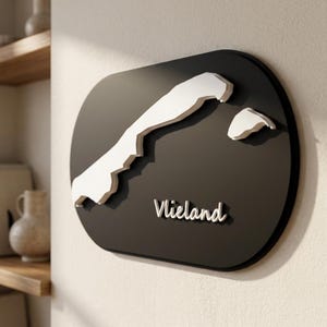 Peut inclure: Une plaque murale ovale noire présentant une carte en relief blanche de l'île de Vlieland. Le texte blanc Vlieland est positionné sous la carte. La plaque a une finition mate lisse et est fixée sur une surface murale de couleur claire.
