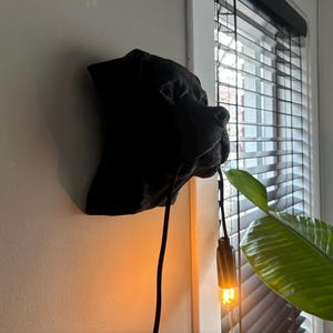 May include: A black dog head wall decor with a light bulb hanging from it. The dog head is mounted on a white wall, with a window and a green plant in the background.