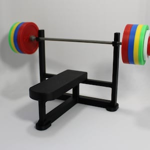 Könnte beinhalten: Eine Miniatur-Hantelbank mit einer Langhantel und bunten Gewichtsscheiben. Die Bank und die Stützen sind schwarz, und die Gewichte sind rot, blau, gelb und grün. Die Langhantel ist silberfarben.