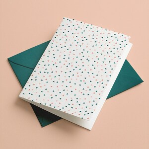 Può includere: Un set di biglietti di auguri e buste. Il biglietto presenta uno sfondo bianco con un motivo di piccoli triangoli rosa e verde acqua. La busta è di colore verde acqua pieno. Il set è esposto su una superficie color pesca chiaro.