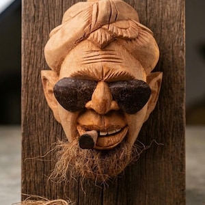 May include: A carved wooden face with sunglasses and a cigar, mounted on a wooden plank. The face has a detailed beard and hair, with a smiling expression. The wood has a natural, rustic appearance.