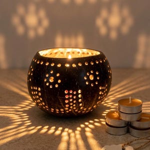 May include: A hand-carved coconut lantern with a lit candle inside, casting patterned shadows. The dark brown lantern has numerous small, circular cutouts. Several tea light candles and a tag that reads "Authentic Handcarved Coconut Lantern" are also visible.