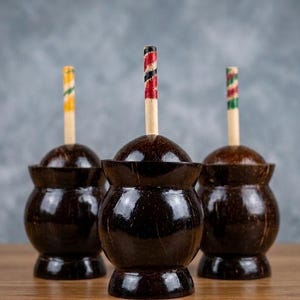 May include: Three dark brown coconut shell containers with decorative wooden tops. Each top has a different colored spiral design: yellow and green, red and black, and green and white. The containers sit on a wooden surface.