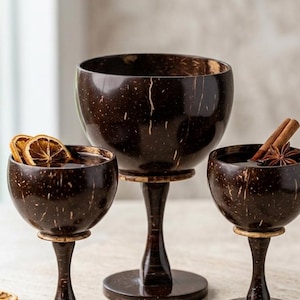May include: Three dark brown coconut shell cocktail glasses with stems. One glass contains dried orange slices, another has a cinnamon stick and star anise. The glasses are on a light-colored surface.