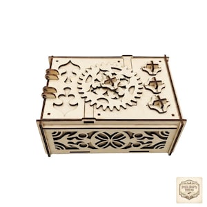 May include: A wooden puzzle box with intricate gear and floral designs. The box features a large gear mechanism on top, with smaller gears and cross-shaped elements. The sides and front of the box have decorative cut-out patterns. The box is made of light-colored wood.
