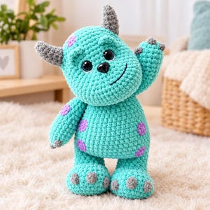 May include: A turquoise crochet monster plush toy featuring purple spots, two grey horns, and grey claws on its hands and feet. The creature has large black plastic eyes, a small black nose, and a stitched smile, with one arm raised in a wave.