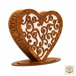 May include: A wooden heart-shaped decorative item with an intricate, swirling design cut out of the front. The item is made of light brown wood with a white background visible through the cutouts. It stands on a matching oval base.