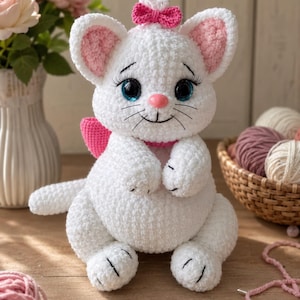 Könnte beinhalten: Ein weißes, gehäkeltes Katzen-Stofftier mit großen blauen Augen, langen schwarzen Wimpern, einer kleinen rosa Nase und gestickten Schnurrhaaren. Die Katze hat rosa Innenohren, eine rosa Schleife auf dem Kopf und ein rosa Halsband mit einer passenden Schleife am Rücken.