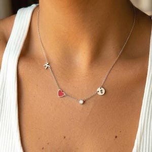 Puede incluir: Un collar de cadena plateada con tres pequeños dijes: una letra K metálica, un colgante en forma de corazón rojo y un disco circular con un grabado de árbol, centrado con una piedra redonda transparente en un engaste plateado.
