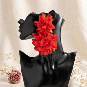 Puede incluir: Un par de pendientes de flores de tela de color naranja vibrante con pétalos superpuestos y fruncidos. Los pendientes se muestran en un busto de maniquí negro, destacando el diseño floral audaz y el color naranja brillante y saturado de la tela.